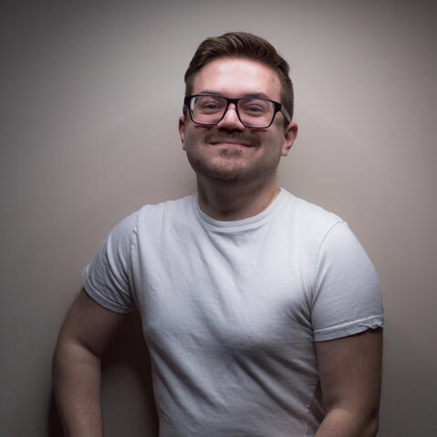 A white man smiles at the camera with a hand resting on his knee.